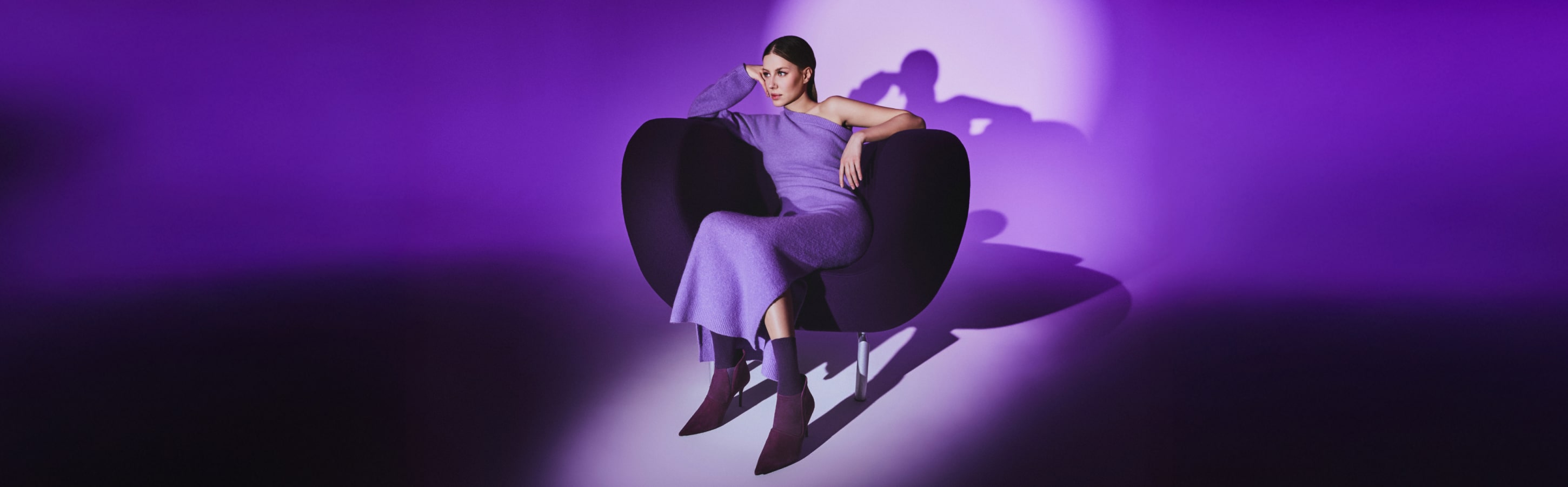 FALKE Cosy Wool Socken – Damenmode in Violett Frau in violettem Strickkleid mit FALKE Cosy Wool Socken, sitzend auf modernem Sessel vor violettem Hintergrund.