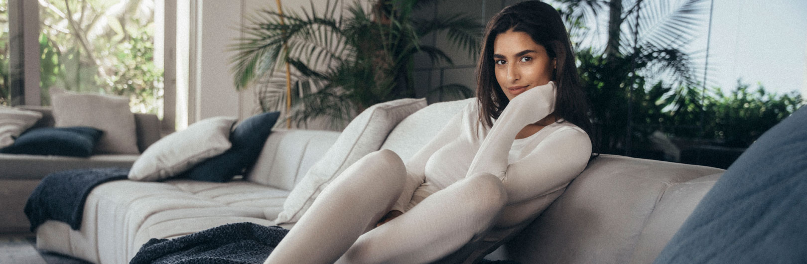 Auf dem Sofa sitzt eine Frau und trägt ein langärmliges Shirt und eine lange Hose aus der ClimaWool Unterwäschen Serie