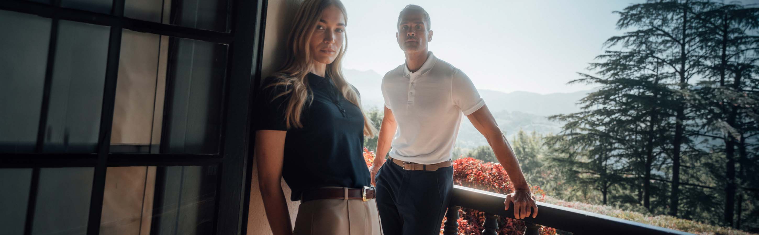 Ein Mann und eine Frau stehen auf einem Balkon, sie trägt ein schwarzes Poloshirt, er trägt ein weisses, sie wenden ihren Blick der Kamera zu