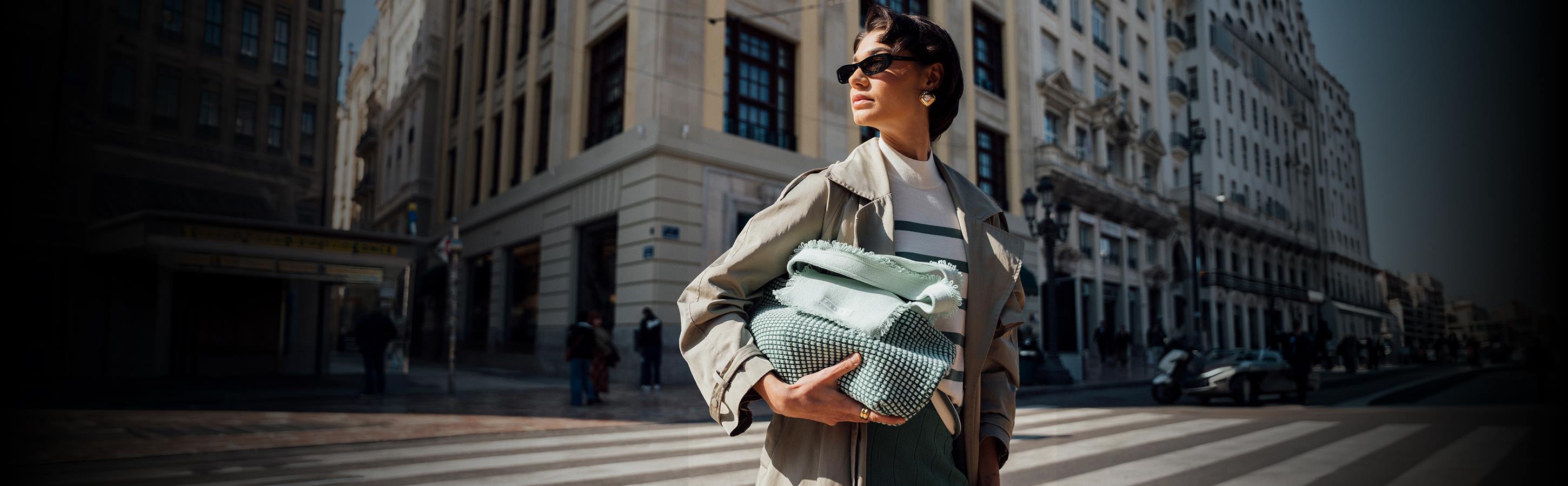 Accessoires für Damen Frau mit Sonnenbrille trägt hellen Mantel und mintgrüne Tasche auf einer sonnigen Straße in urbanem Umfeld.