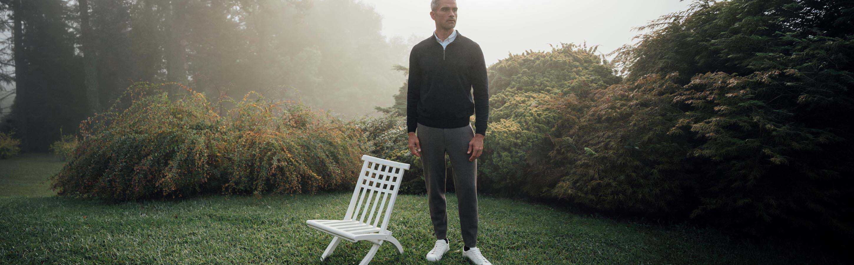 Lambswool Pullover für Herren Mann steht im Garten mit schwarzem Pullover, grauer Hose und weißen Sneakern.