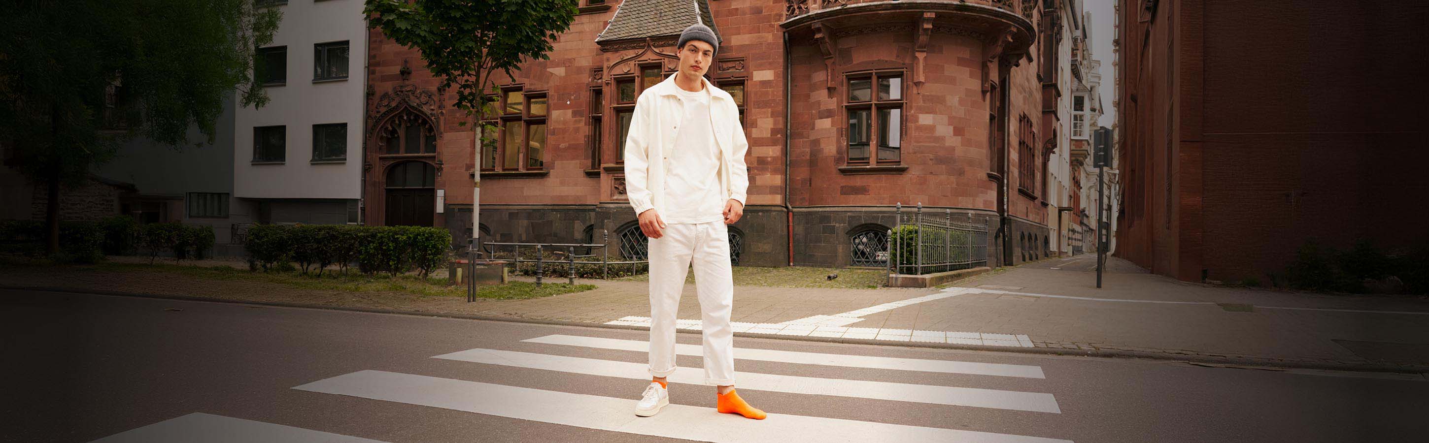 Mann trägt orangefarbene Sneakersocken der Serie Cool Kick von FALKE und steht auf einem Zebrastreifen auf der Straße