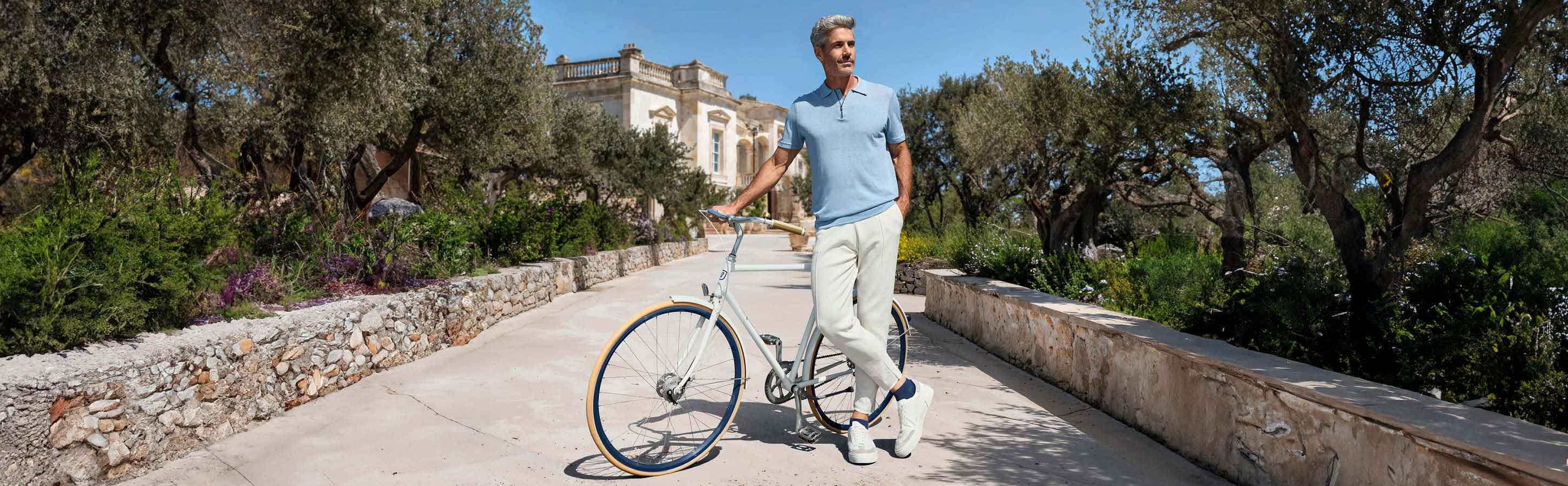 Mann trägt dunkelblau Sneakersocken von FALKE mit weißen Sneakern, einer weißen Hose und einen hellblauen Poloshirt und steht mit einem Fahrrad auf einem Weg im Süden