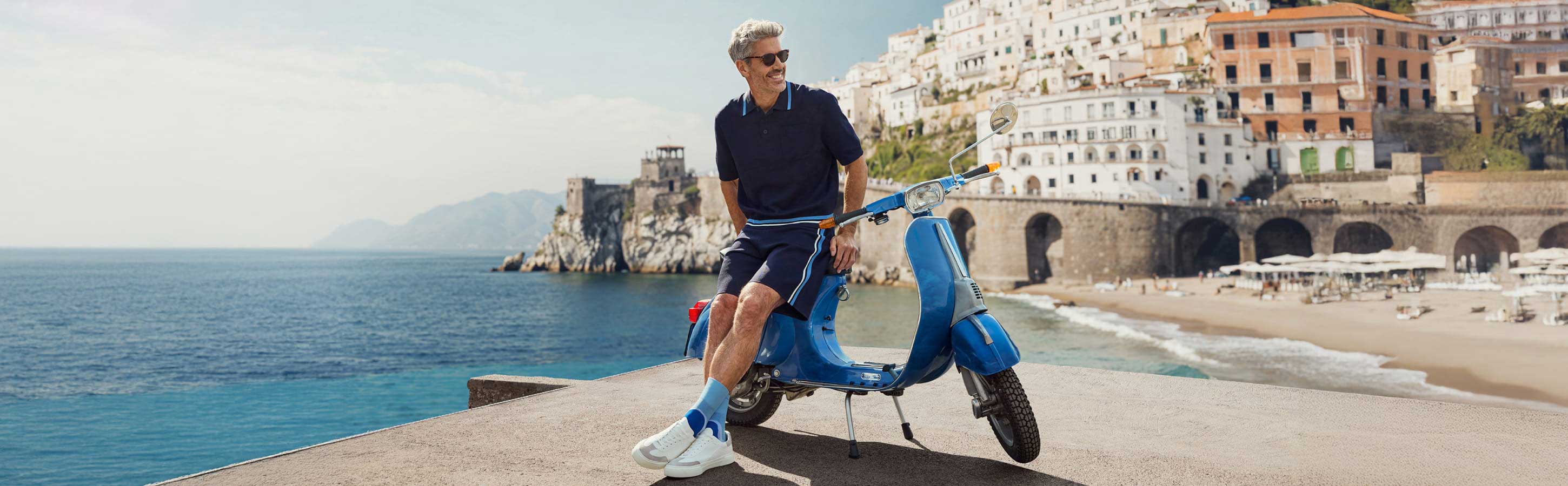 Mann trägt blaue Socken von FALKE und blaue Freizeitkleidung und lehnt sich an einen Motorroller, der am Meer steht