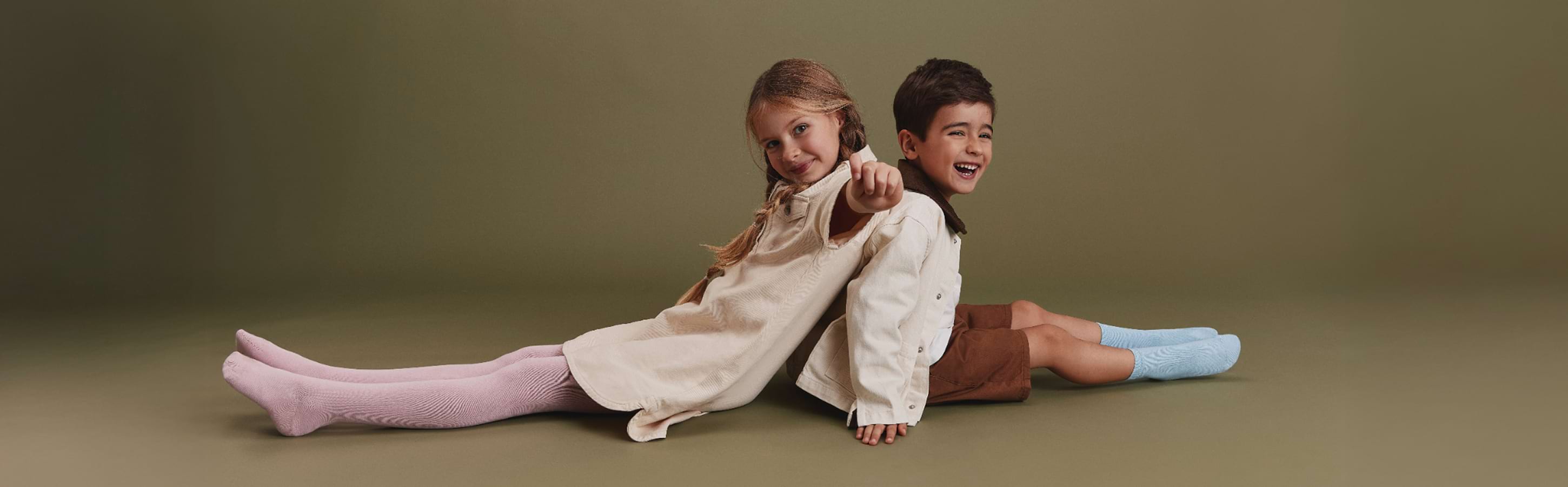 Zwei Kinder sitzen Rücken an Rücken vor neutralem Hintergrund, lachen und tragen die FALKE Family Styles; das Mädchen trägt eine helle FALKE Strumpfhose, der Junge farbige FALKE Socken zu schlichten Outfits