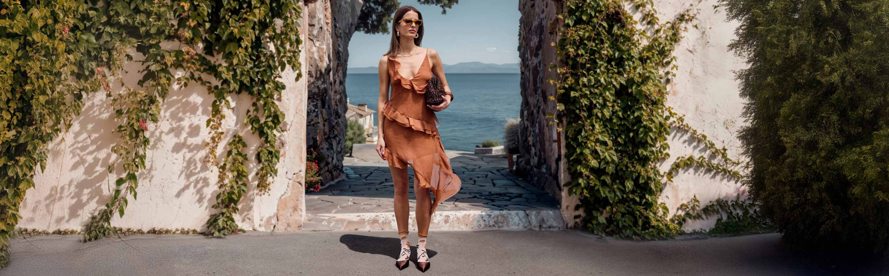 Frau im Sommerkleid geht durch einen mediterranen Durchgang mit Meerblick und trägt gemusterte FALKE Socken zu spitzen Schuhen