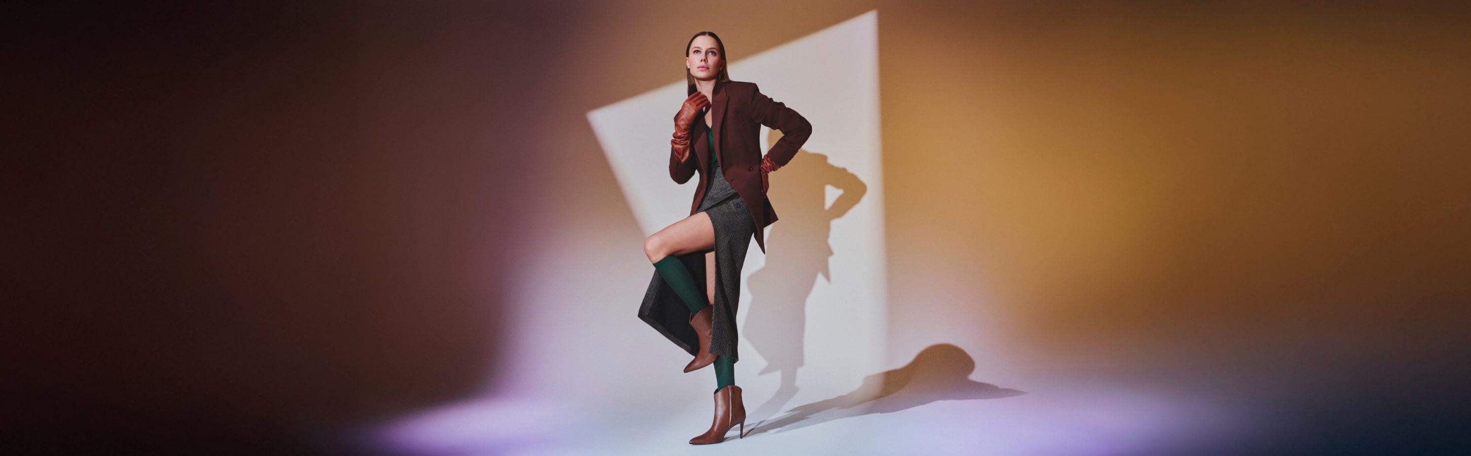FALKE Colour Competence – Stilvolle Damen mit Clima Wool Kniestrümpfen Frau in eleganter Herbstmode trägt grüne FALKE Clima Wool Kniestrümpfe, kombiniert mit Rock, Blazer und Stiefeln vor farbigem Hintergrund