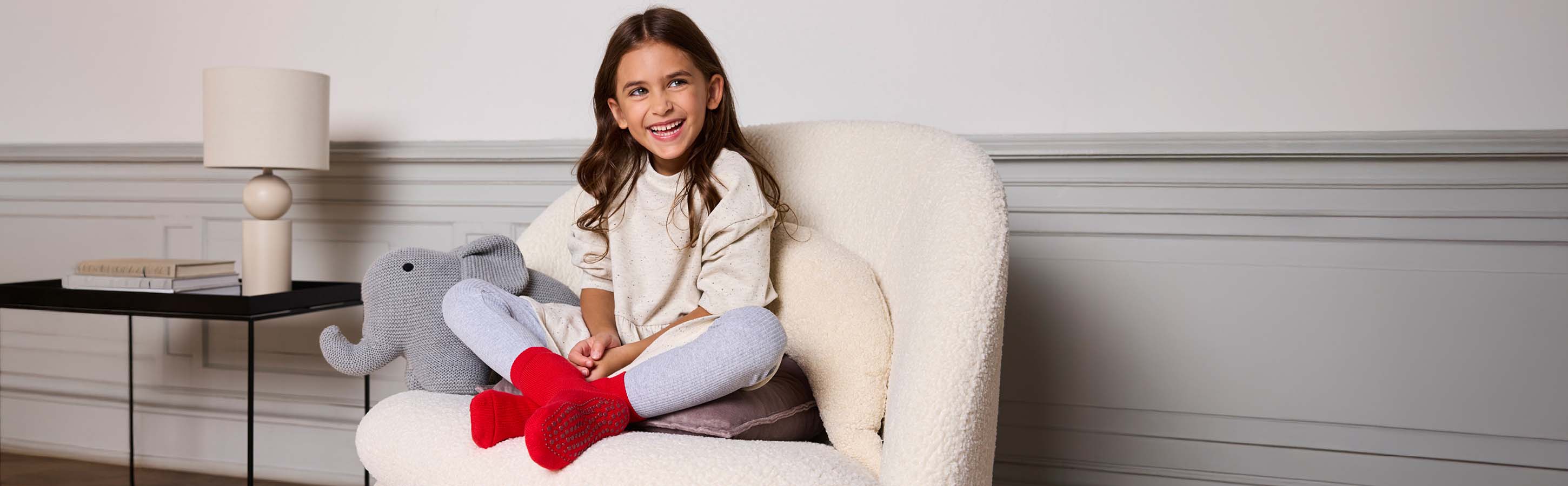 Lachendes Mädchen trägt rote FALKE Catspads Hausstrümpfe und graue Leggings auf einem Sessel – gemütlicher Homewear-Look für Kinder