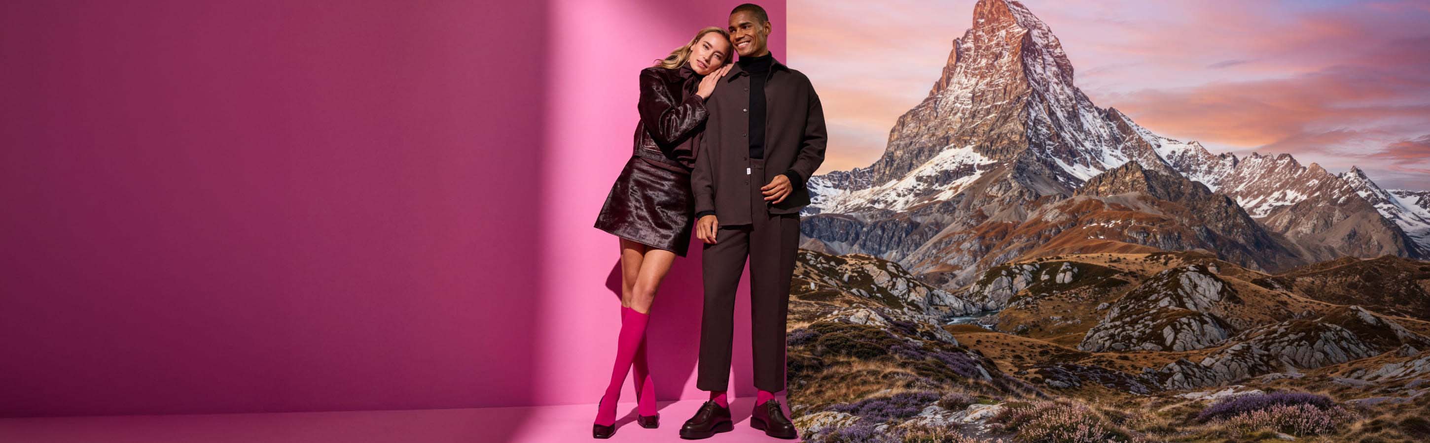 Die neue Kollektion von FALKE Ein Paar steht vor einer farbigen Wand und einem begrünten Berg, sie tragen elegante Kleidung und sie lehnt sich an ihn