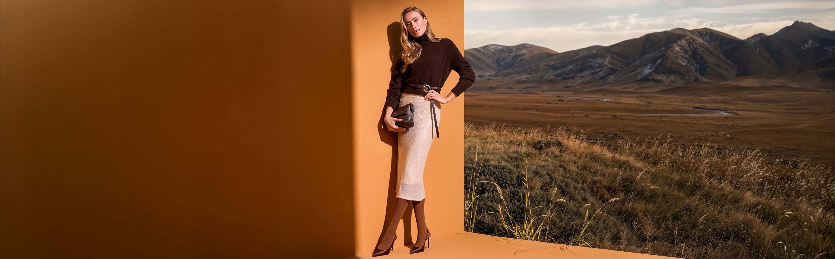 FALKE Pure Matt 50 blickdichte Feinstrumpfhose Frau trägt die braune FALKE Pure Matt 50 Feinstrumpfhose zu Rollkragenpullover, Paillettenrock und Pumps vor herbstlicher Berglandschaft